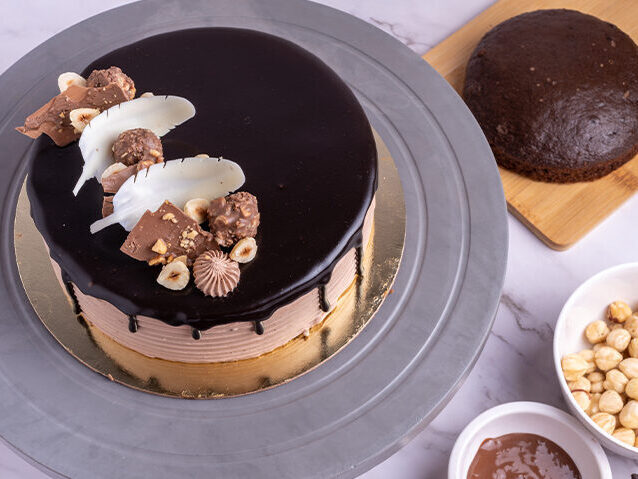 A chocolate glazed cake, decorated with assorted chocolates and white chocolate pieces, presented on a grey stand, with ingredients displayed around it.